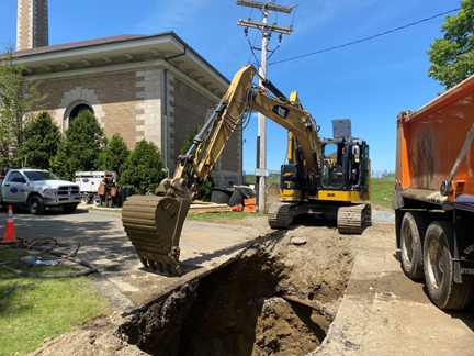 An excavator digs out a deep pit that crosses what looks like a driveway.