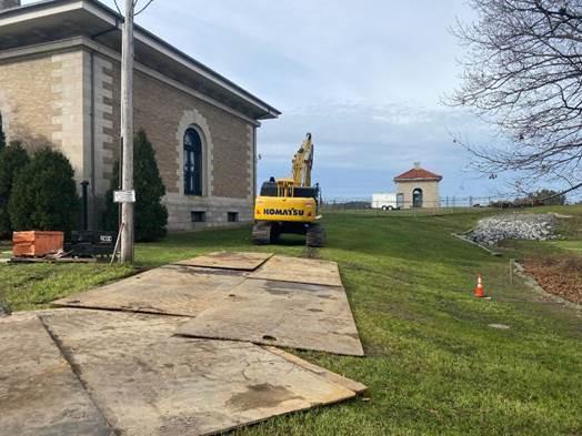On a small grassy slope behind a building, an excavator drives across a plywood path, onto the grass.