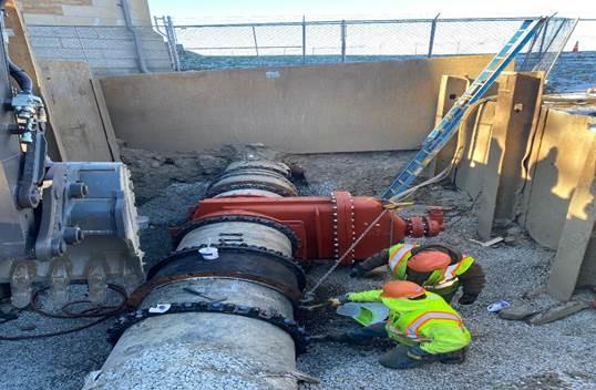 In a shallow pit, to workers are crouched looking at a large pipe.