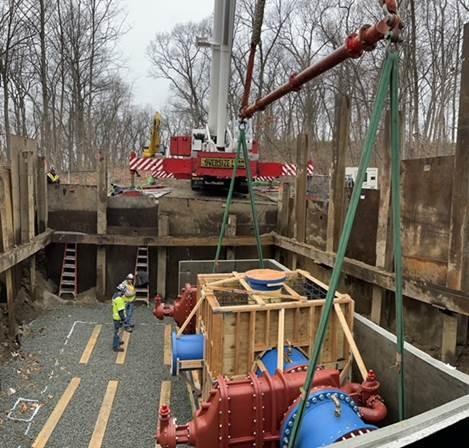 In a large pit, workers watch as a large valve gets lowered in. The pit looks to be around 15 feet deep and twice as wide.
