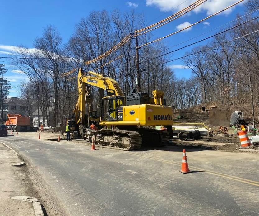 On a street near a forested area, an excavator and several workers work on tearing up a section of street.