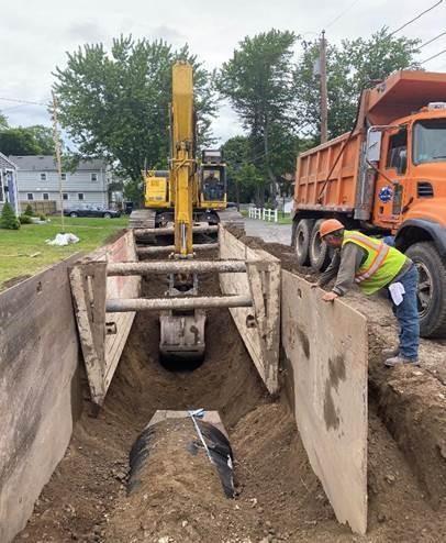 An excavator digs out a trench while a working installs wood supports along the side.