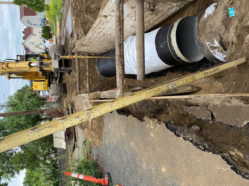 A trench in a residential street, with a large black pipe in it. A crane is attached to the pipe.