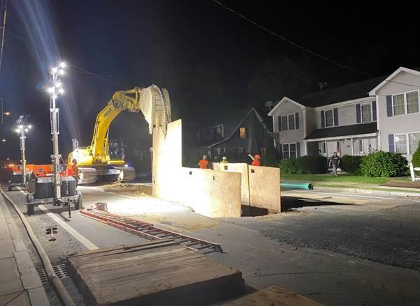 On a residential street at night, an excavator sits over a hole with ply wood supporting the sides of it.