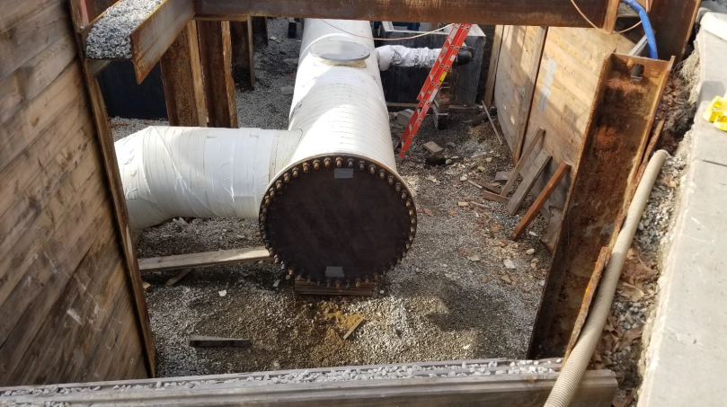 In a trench, there is the end of a large white pipe. Before the pipe ends, there is another pipe branching off it.