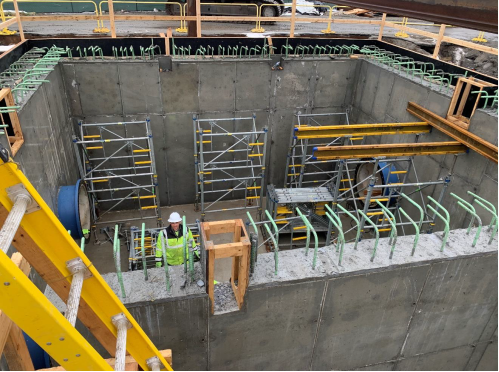 A man stands at the bottom on a large, concrete sided pit in the ground. The pit seems to be about twice his height, and green support beams are seen coming out of the concrete.