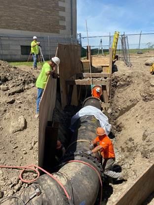In a pit, two works work on a pipe section, while a worker looks on over the support wall.