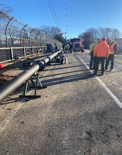 A long pipe is propped up, running down the side of a road. Works stand around on the road.