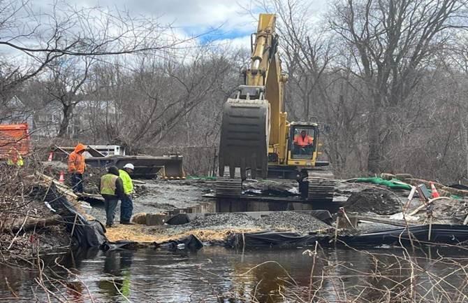 Near a creek, several workers stand around an excavator reaching toward the river.