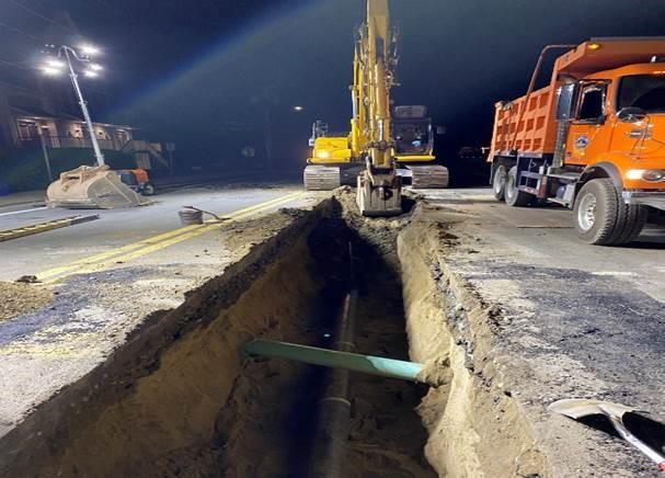 A 3 foot wide trench in the street. A black pipe runs along the bottom, toward an excavator extending the trench.