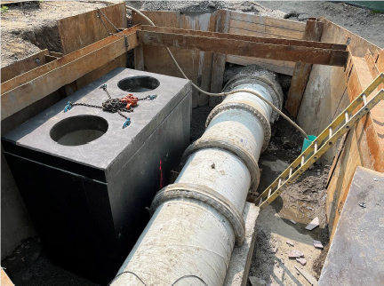 In a pit in the ground a large pipe runs next too a large mental box.