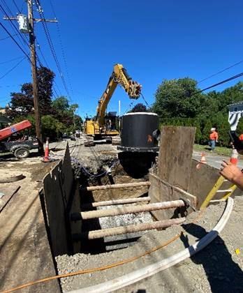 A crane lifts a large black cylinder toward a square pit in the street.
