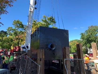 A large metal cube object is lowered into a pit in the ground by a crane.