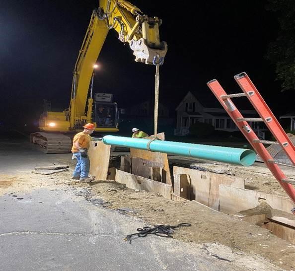 An crane lowers a long blue pipe into a trench while a worker guides it.