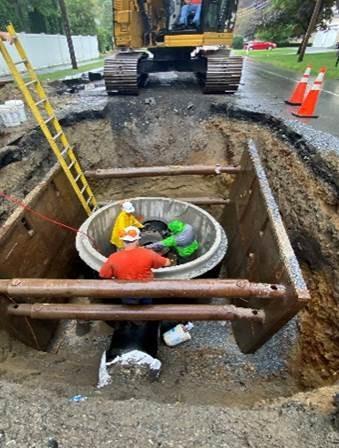 In a square pit, about as deep as the workers, two men work on a cylinder that has just been place there.