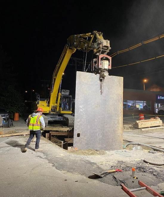 A crane lowers a tall sheet of metal into a trench in the street.
