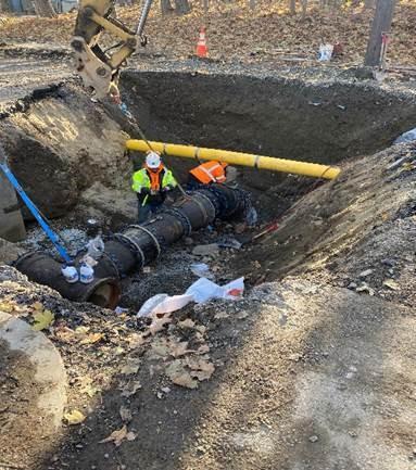 In a large pit, men work on attaching a chain to a large black pipe.