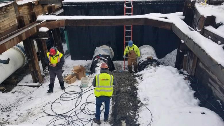 Three workers stand in a lightly snow covered pit in the ground.
