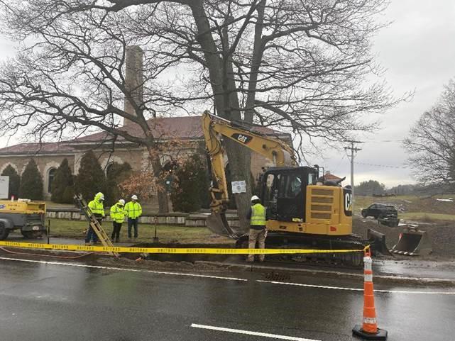 On a residential street, 4 workers in high visibility clothing stand around a trench being excavated.