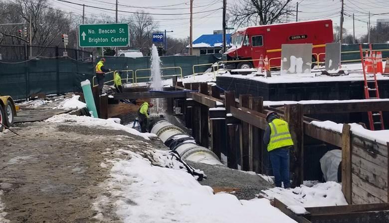 A large white pipe runs the length of a narrow trench. Workers are in and above the trench. At the far end, what spurts out.
