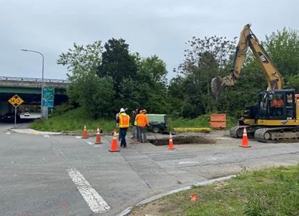 At an intersection, and excavator digs a small pit while workers standby.