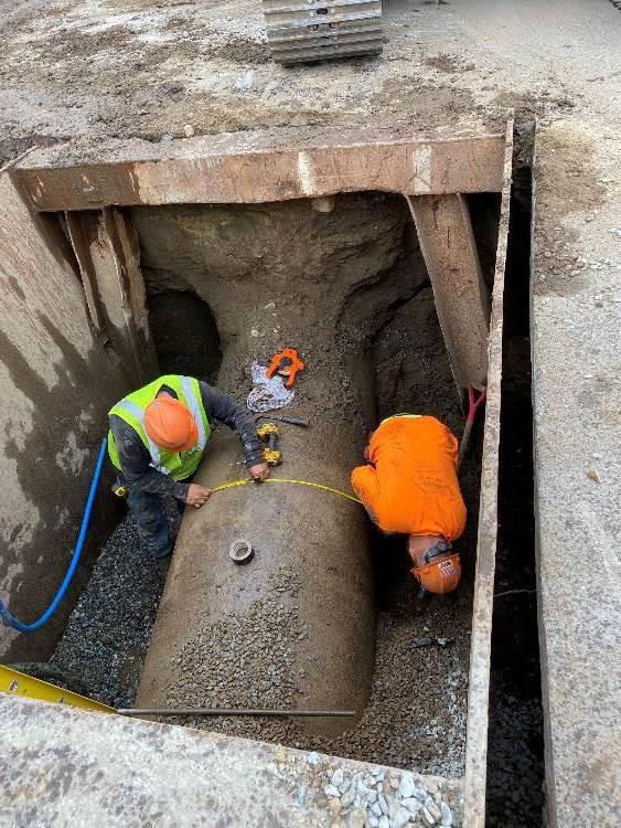 In a pit two workers measure the circumference of a pipe.