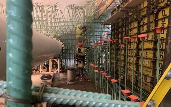 In a large, covered pit two workers install rebar for the concrete walls.