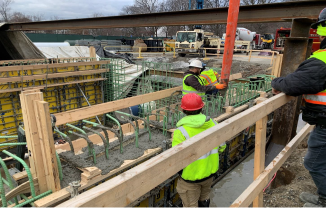 The top of the concrete support walls of a pit. Workers work over the rebar coming out of the concrete.