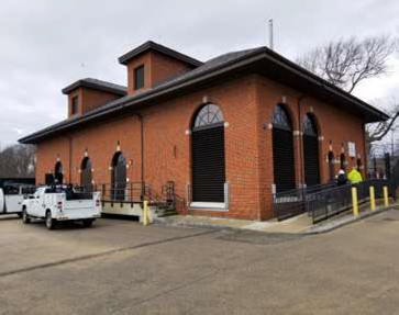 A brown brick building with a dark trims. The windows are arched and several yellow concrete pillars circle the building.