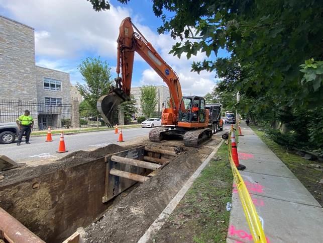 An orange digger sits on the edge of a square pit dug on a city street. 