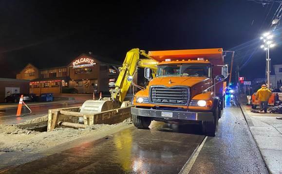 A orange dump truck next to a hole dug in the street. Its night and the area is lit by large flood lights.