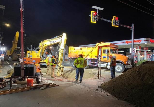 Under traffic lights at a intersection next to a gas station, workers stand around equipment digging a hole, lit by flood light