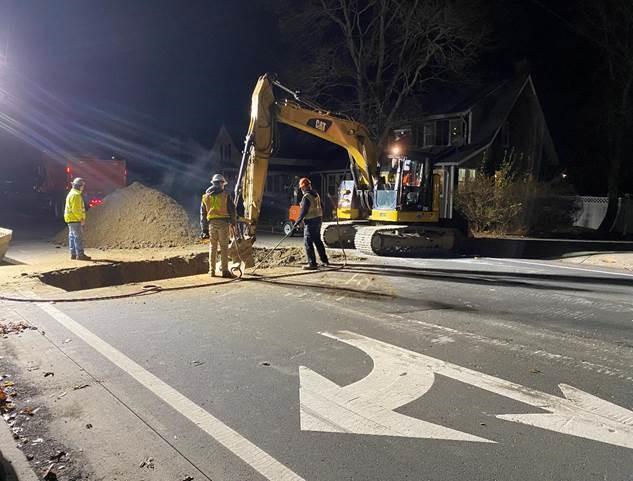Behind turn lane arrows, an excavator digs a hole, while 3 crew stand watching.